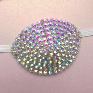 May include: An eye patch adorned with iridescent rhinestones, secured by a white elastic band. The rhinestones shimmer with multiple colours, creating a dazzling effect. The eye patch is designed to cover one eye.