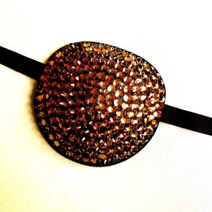 May include: An oval eye patch covered in numerous small, reflective bronze-coloured crystals. The patch is attached to a thin, smooth black elastic strap that extends horizontally from both sides of the central oval piece.