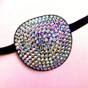 May include: An oval-shaped black eye patch, densely covered in iridescent rhinestones. The rhinestones reflect a spectrum of colours. It has a black strap for securing. The eye patch is set against a pink background.