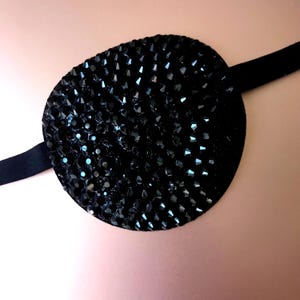 May include: A black eye patch with a round, black, rhinestone-covered patch. The patch is attached to a black elastic band.
