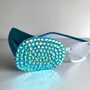 May include: A teal eye patch with an oval, bedazzled design, combined with clear-framed spectacles. The patch is covered in small, sparkling, turquoise-coloured rhinestones. The spectacles have thin, blue arms.