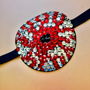 May include: An oval eye patch featuring a dense arrangement of sparkling red and clear faceted rhinestones. The centre of the patch contains a cluster of dark black stones, and the piece is attached to a simple, smooth black elastic strap.