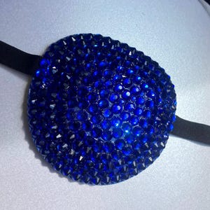 May include: A circular eye patch covered in small, reflective royal blue rhinestones. The patch features a smooth, rounded surface texture created by the dense arrangement of faceted gems and is attached to a thin, black elastic strap on each side.