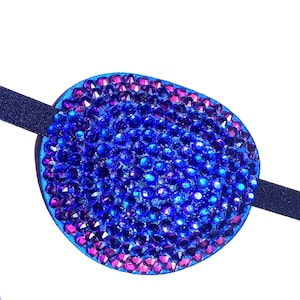 May include: An oval eye patch covered in vibrant blue and purple faceted rhinestones. The patch features a dense arrangement of sparkling gems that create a textured surface. A dark elastic strap is attached to the sides for secure placement.