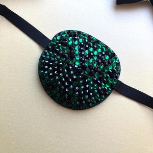 May include: An eye patch featuring a circular base covered in a dense arrangement of faceted black and emerald green crystals. The patch is attached to a thin, flexible black elastic strap designed to secure the piece around the head.