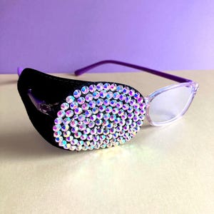 May include: Eyeglasses with a black eye patch adorned with iridescent rhinestones. The glasses feature a purple frame and clear lenses. The background is a light purple.