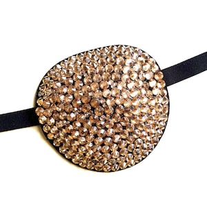 May include: An eye patch with a black base, densely covered in amber-toned rhinestones. The oval-shaped patch is attached to a black elastic strap. This accessory is designed for fashion or costume purposes, adding a touch of glamour.