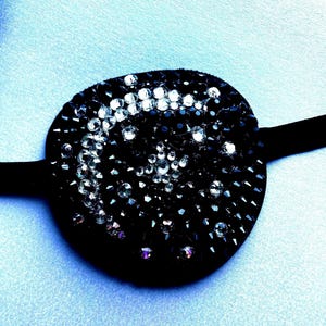 May include: A circular black eye patch featuring a crescent moon and star design created with clear and black crystals. The patch is attached to a black fabric strap and is heavily embellished with dark studs and faceted rhinestones.