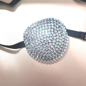 May include: A circular eye patch covered in numerous clear, faceted rhinestones that reflect light. The patch is attached to a thin, black elastic strap designed to secure the item in place. The base of the patch appears to be a soft pink fabric.