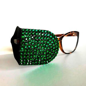 May include: A black eye patch adorned with many green rhinestones, alongside tortoiseshell spectacles. The patch is designed to cover one eye, providing a fashionable and decorative accessory.