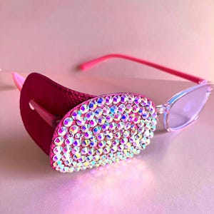May include: Cat-eye sunglasses with a pink frame and a magenta leather eye patch. The eye patch is covered in iridescent rhinestones. The lenses are a light, translucent colour. The background is a soft pink.