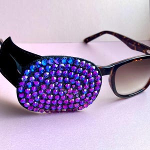 May include: Sunglasses with a decorative eye patch. The patch is oval and adorned with blue and purple rhinestones. The sunglasses feature a tortoiseshell frame and brown lenses. The background is a pale pink.