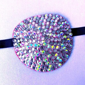 May include: An oval eye patch covered in iridescent, multi-faceted clear rhinestones that reflect various colours. The patch is mounted on a thin, black elastic strap that extends from both sides of the circular, pink-edged fabric base.