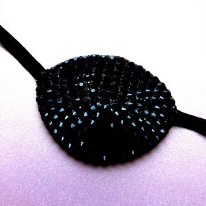 May include: An oval-shaped eye patch covered in small, faceted black crystals that create a textured, reflective surface. The patch is attached to a thin, black elastic strap that extends from both sides, designed to secure the piece in place.