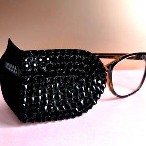 May include: Black eye patch covered in small, sparkling black gems, attached to a pair of eyeglasses with a tortoiseshell frame. The patch is designed to cover one eye, with a curved shape.