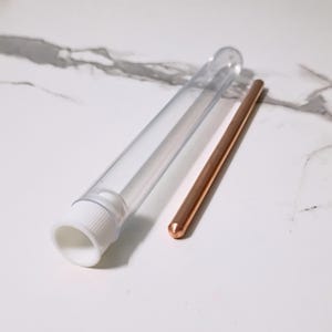 May include: A clear plastic tube with a white cap and a copper-colored rod. The tube is open, and the rod is lying next to it. The background is a white surface with gray veining.
