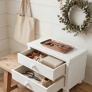 May include: A white wooden two-drawer chest with round knobs, featuring an open top drawer containing wooden rings, twine, and a book, and an open bottom drawer with scissors and twine. A copper tray with keys and glasses sits on top.