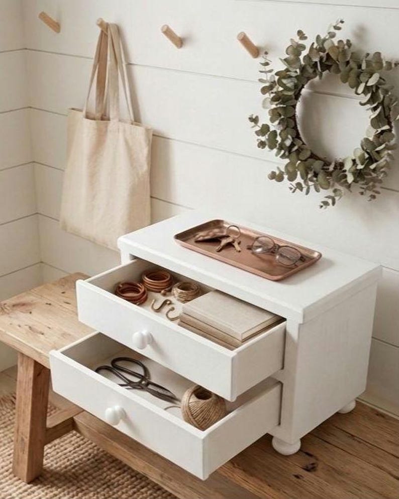 May include: A white wooden two-drawer chest with round knobs, featuring an open top drawer containing wooden rings, twine, and a book, and an open bottom drawer with scissors and twine. A copper tray with keys and glasses sits on top.