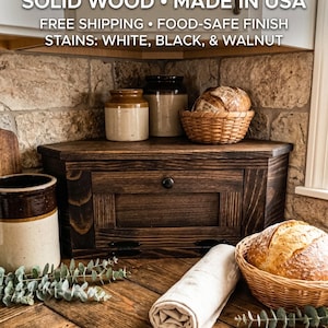 May include: A dark walnut-stained wooden bread box with a single drawer and a round black knob. The box is on a wooden surface with a basket of bread, a rolled-up cloth, and ceramic jars. The text "SOLID WOOD • MADE IN USA" is visible.