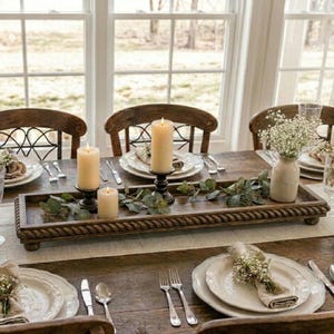 May include: A rectangular wooden tray with a rope-textured border, featuring three cream-colored pillar candles and green eucalyptus sprigs. The tray is placed on a dining table set with white plates, silverware, and small vases of baby's breath.