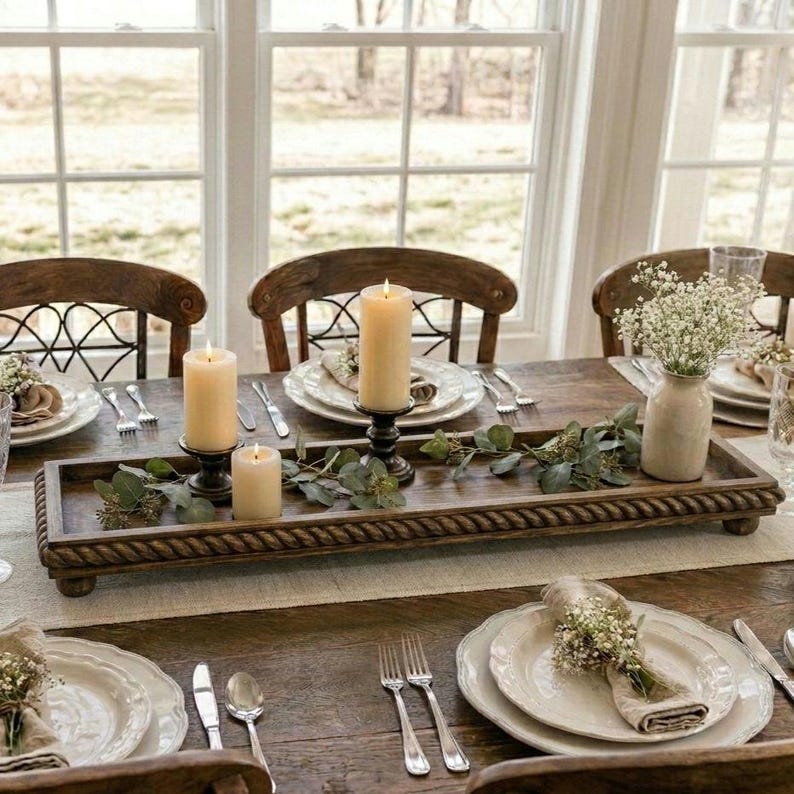 May include: A rectangular wooden tray with a rope-textured border, featuring three cream-colored pillar candles and green eucalyptus sprigs. The tray is placed on a dining table set with white plates, silverware, and small vases of baby's breath.