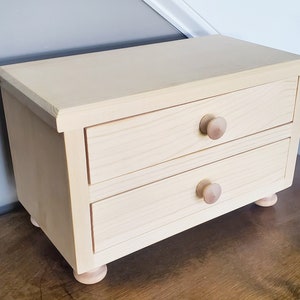 K&ouml;nnte beinhalten: Eine kleine Holzkiste mit zwei Schubladen und gedrehten Holzf&uuml;&szlig;en. Die Kiste ist in einer hellen Holzfarbe lackiert und hat ein schlichtes Design.