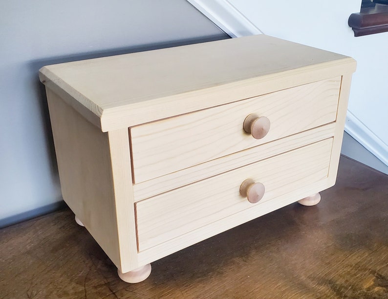 K&ouml;nnte beinhalten: Eine kleine Holzkiste mit zwei Schubladen und gedrehten Holzf&uuml;&szlig;en. Die Kiste ist in einer hellen Holzfarbe lackiert und hat ein schlichtes Design.