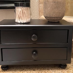 May include: A black wooden desktop organizer featuring two horizontal drawers with round knobs, supported by four small bun feet. A clear glass jar filled with white cotton swabs and a textured beige cylindrical container sit on top.