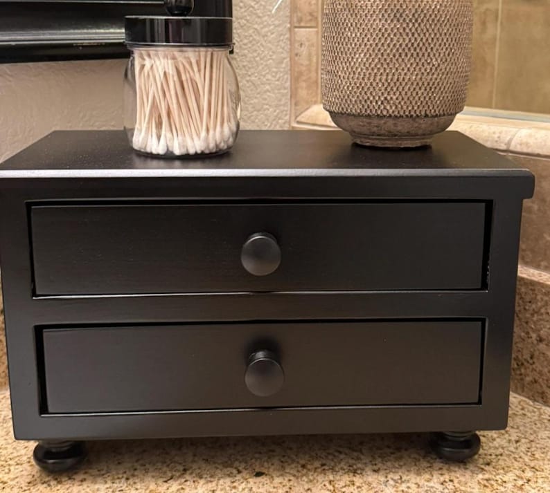 May include: A black wooden desktop organizer featuring two horizontal drawers with round knobs, supported by four small bun feet. A clear glass jar filled with white cotton swabs and a textured beige cylindrical container sit on top.