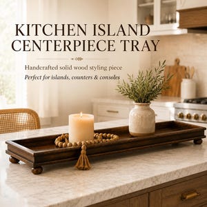 May include: A rectangular dark brown wooden tray with four small ball feet. The tray holds a white pillar candle, a string of wooden beads with a tassel, and a small ceramic vase containing green foliage. The text reads KITCHEN ISLAND CENTERPIECE TRAY.