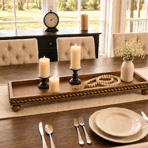 May include: A rectangular brown wooden tray with a rope-style border and ball feet. The tray holds three cream-colored pillar candles in black holders, a string of wooden beads with a tassel, and a small white vase containing white flowers.