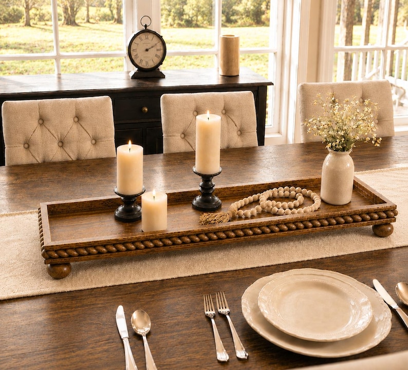 May include: A rectangular brown wooden tray with a rope-style border and ball feet. The tray holds three cream-colored pillar candles in black holders, a string of wooden beads with a tassel, and a small white vase containing white flowers.