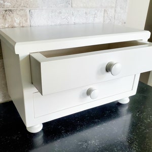 May include: A white wooden desktop organizer featuring two drawers with round knobs. The top drawer is partially extended, revealing an open interior. The unit is supported by four small, rounded feet, providing a stable base for this storage piece.