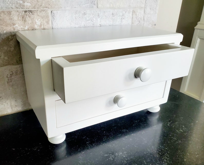May include: A white wooden desktop organizer featuring two drawers with round knobs. The top drawer is partially extended, revealing an open interior. The unit is supported by four small, rounded feet, providing a stable base for this storage piece.