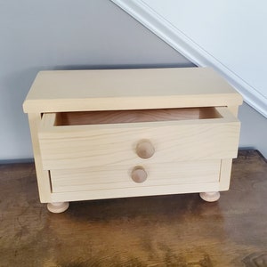 K&ouml;nnte beinhalten: Eine kleine Holzkiste mit zwei Schubladen und runden Holzgriffen. Die obere Schublade ist ge&ouml;ffnet und zeigt den leeren Innenraum.
