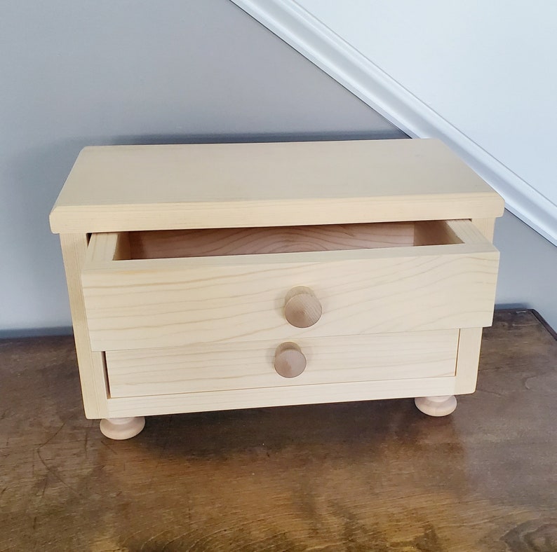 K&ouml;nnte beinhalten: Eine kleine Holzkiste mit zwei Schubladen und runden Holzgriffen. Die obere Schublade ist ge&ouml;ffnet und zeigt den leeren Innenraum.