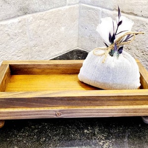 Pode incluir: Uma bandeja retangular de madeira com interior castanho claro e moldura castanho escuro, apoiada em pequenos p&eacute;s arredondados. Uma pequena bolsa de tecido branco amarrada com barbante e decorada com flores secas est&aacute; dentro da bandeja.