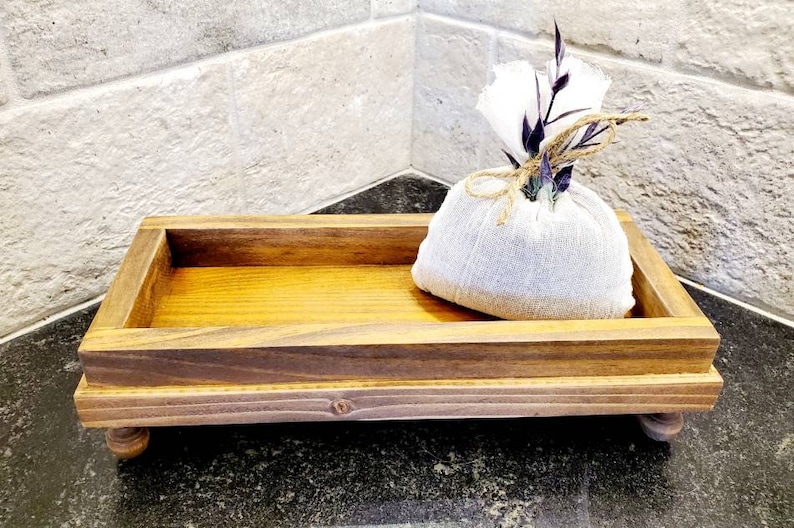 Pode incluir: Uma bandeja retangular de madeira com interior castanho claro e moldura castanho escuro, apoiada em pequenos p&eacute;s arredondados. Uma pequena bolsa de tecido branco amarrada com barbante e decorada com flores secas est&aacute; dentro da bandeja.