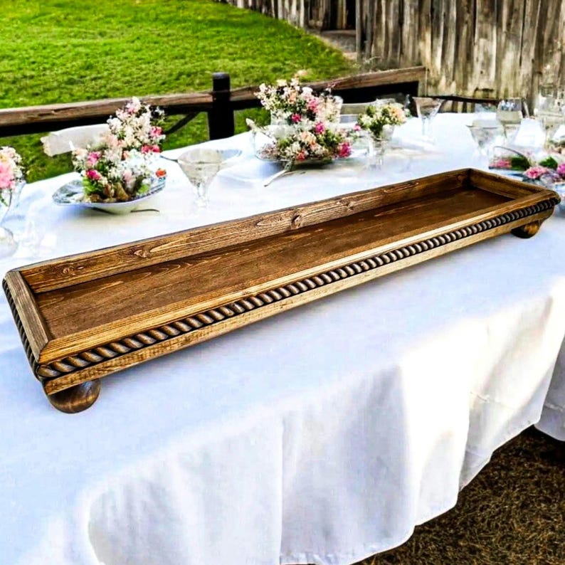May include: A long, rectangular wooden tray with a dark brown stained finish. The tray features a decorative rope-style carved border along the edges and is supported by four small, rounded feet at the corners, providing a raised platform.