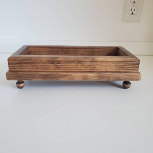 Pode incluir: Uma bandeja retangular de madeira com mancha castanha escura. A bandeja &eacute; elevada por quatro pequenos p&eacute;s redondos de cor bronze. O gr&atilde;o da madeira &eacute; vis&iacute;vel, adicionando textura. O design da bandeja &eacute; simples e funcional.