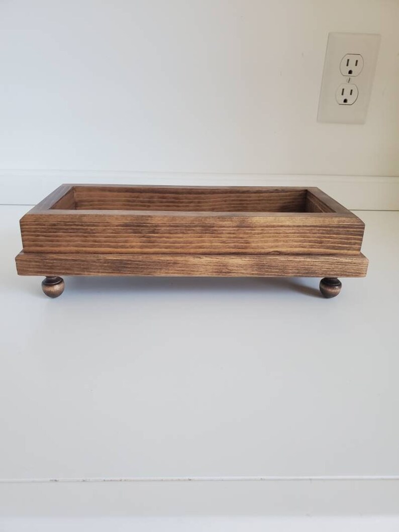 Pode incluir: Uma bandeja retangular de madeira com mancha castanha escura. A bandeja &eacute; elevada por quatro pequenos p&eacute;s redondos de cor bronze. O gr&atilde;o da madeira &eacute; vis&iacute;vel, adicionando textura. O design da bandeja &eacute; simples e funcional.