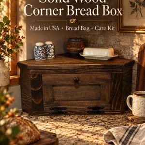 May include: A dark brown solid wood corner bread box featuring a front-hinged door with a small round knob. The text on the image reads: Solid Wood Corner Bread Box, Made in USA, Bread Bag + Care Kit. The box has a triangular shape.