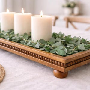 May include: A rectangular wooden tray with a beaded edge and four rounded feet, filled with green eucalyptus leaves and three white pillar candles. The tray is constructed from medium-toned wood and serves as a base for the greenery and candles.