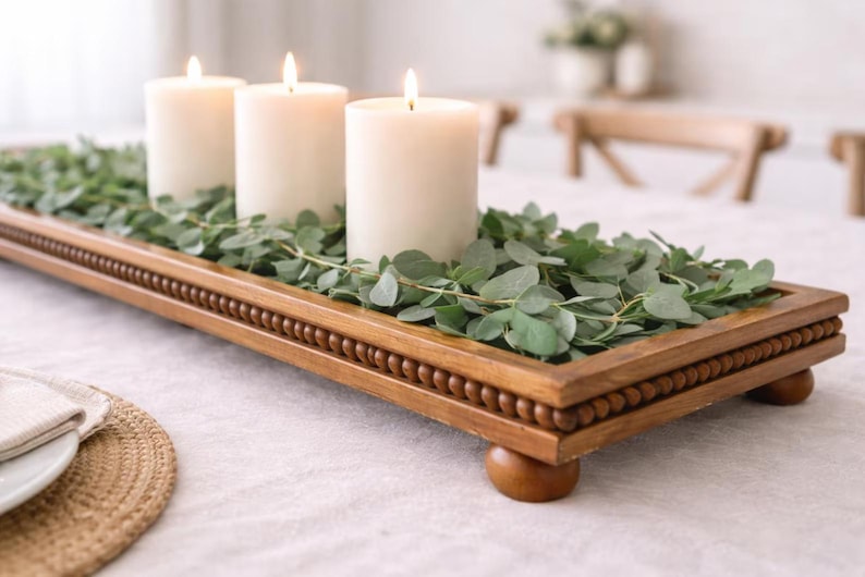 May include: A rectangular wooden tray with a beaded edge and four rounded feet, filled with green eucalyptus leaves and three white pillar candles. The tray is constructed from medium-toned wood and serves as a base for the greenery and candles.