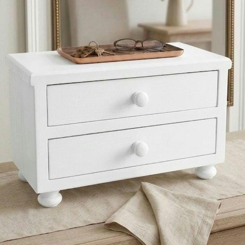 May include: A white wooden desktop organizer featuring two horizontal drawers with round knobs, supported by four rounded feet. A rectangular wooden tray sits on top, holding a pair of dark-rimmed eyeglasses and a metal key ring with keys.