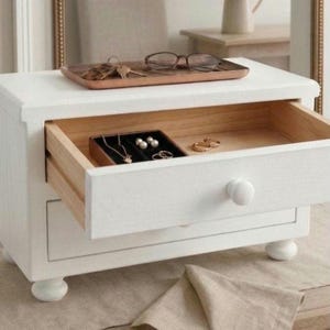 May include: A white wooden jewelry box with two drawers and four rounded feet. The top drawer is open, revealing a black velvet insert holding a gold necklace, pearl earrings, and rings. A wooden tray with eyeglasses and keys sits on top.