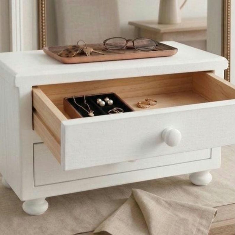 May include: A white wooden jewelry box with two drawers and four rounded feet. The top drawer is open, revealing a black velvet insert holding a gold necklace, pearl earrings, and rings. A wooden tray with eyeglasses and keys sits on top.