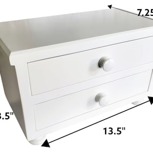 May include: A white wooden desktop organizer featuring two horizontal drawers with round knobs. The unit measures 13.5 inches in width, 8.5 inches in height, and 7.25 inches in depth, as indicated by the black dimension arrows on the image.