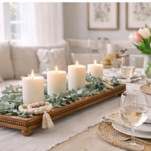May include: A rectangular wooden tray with beaded edges and four small feet, holding four white pillar candles, green eucalyptus foliage, and a strand of light-colored wooden beads with a tassel, arranged as a centerpiece on a dining table.