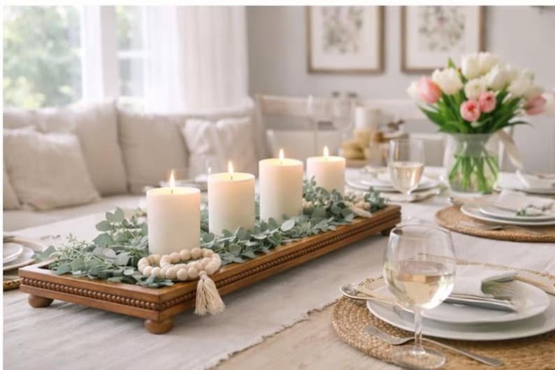 May include: A rectangular wooden tray with beaded edges and four small feet, holding four white pillar candles, green eucalyptus foliage, and a strand of light-colored wooden beads with a tassel, arranged as a centerpiece on a dining table.